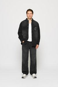 Black corduroy shirt with front pocket and embroidered detail, paired with matching black corduroy pants and white athletic shoes.