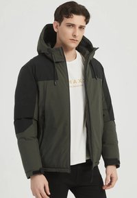 Veste rembourrée verte avec une section supérieure noire, capuche ajustable, poches zippées et une texture de tissu lisse. Portée sur une chemise blanche.
