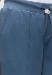 Κοντινό πλάνο μπλε φόρμας με ριμπ λάστιχο στη μέση, τσέπη στην πλαϊνή ραφή και σκούρο κορδόνι με διαφανές πλαστικό στοπ.