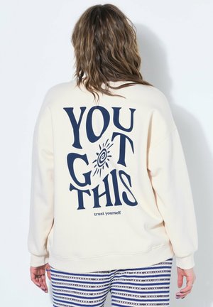 Osoba z falującymi włosami nosi kremową bluzę z granatowym napisem "YOU GOT THIS" oraz "zaufaj sobie", zestawioną z pasiastymi niebiesko-białymi spodniami.