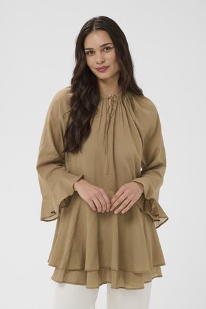 Moteris su ilgais tamsiais plaukais, vilkinti laisvą, sluoksniuotą rudos spalvos palaidinę su raišteliais ties kaklu ir išplatėjusiais rankogaliais, stovinti prie paprasto pilko fono.