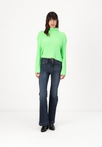 Pulover verde cu rigle, cu un decolteu înalt, asortat cu blugi evazați de culoare albastru închis. Modelul poartă cizme negre până la gleznă și stă în fața unui fundal alb.