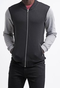 Černá bomber bunda na zip se šedými rukávy, žebrovaným límcem a manžetami, vyrobená z měkké látky. Má boční kapsy a hladký zip.