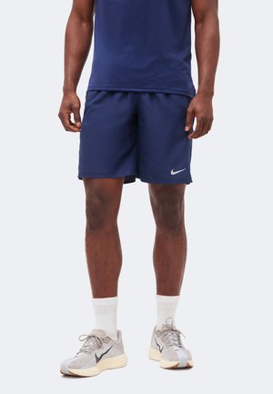 Mand iført marineblå sportsshorts med hvidt Nike-logo, hvide mellemlange sokker og lysegrå løbesko, stående på en ensfarvet baggrund.