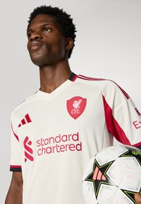 Kreemikatsega spordisärk punaste aksentide ja logoga, millel on lühikesed varrukad ja V-kaelus. Modelle hoiab käes valget ja musta jalgpalli.