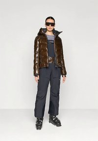 Doudoune en tissu marron brillant avec capuche, associée à un pantalon de ski bleu foncé avec poches. Le modèle porte des chaussures de ski et des lunettes de soleil.