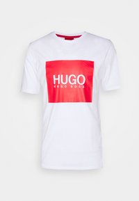 Bílá bavlněná tričko s výrazným červeným obdélníkovým grafikou, na které je bíle napsáno "HUGO" a "HUGO BOSS". Krátké rukávy a kulatý výstřih.