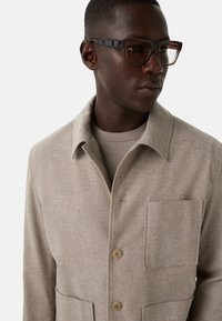 Mand iført skildpaddeformede rektangulære briller og en beige uld skjortejakke over en matchende beige skjorte, der kigger til højre.