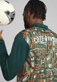 Zelená bunda s dlhými rukávmi a textúrovaným hnedo-bielym vzorovaným chrbtom, na ktorom je bielym písmom napísané "CÔTE D'IVOIRE". Futbalová lopta v popredí.
