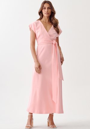 Pink wrap dress with a deep v-neck, short sleeves, and a tied waist. Smooth, lightweight fabric falls to ankle length. Clear heeled sandals.