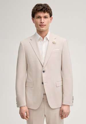 Jeune homme aux cheveux bouclés portant un costume beige et une chemise blanche, face à un fond clair uni.