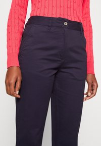 Detailní záběr na osobu oblečenou v tmavě modrých kalhotách a jasně růžovém svetru s pleteným vzorem a dlouhým rukávem, s uvolněnýma rukama po stranách.