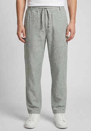 Mann trägt hellgraue Leinenhose mit Kordelzug und weißen Sneakers, steht vor einem einfarbigen hellgrauen Hintergrund.