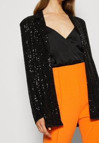 Blazer negro con lentejuelas y diseño estructurado, de mangas largas, superpuesto sobre un top de satén negro y combinado con pantalones sastre de color naranja brillante.