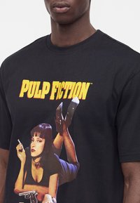 Μαύρο μπλουζάκι με κίτρινο κείμενο "Pulp Fiction" και μια εικόνα με μια γυναίκα ξαπλωμένη που κρατάει τσιγάρο και όπλο στο μπροστινό μέρος.