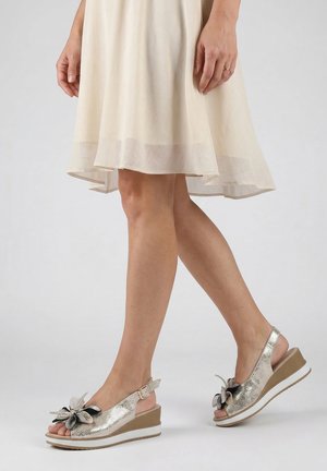 Vrouw draagt een licht beige jurk en metalen sleehak-sandalen met bloemenversiering op de tenen tegen een effen achtergrond.