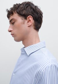 Camicia a righe blu e bianche realizzata in tessuto leggero, con colletto strutturato e chiusura a bottoni. Cuciture visibili lungo le cuciture.