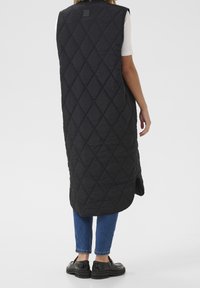 Długi, czarny, pikowany kamizelka w diamentowy wzór, z otwartym przodem i bocznymi rozcięciami, połączona z ciemnymi dżinsami i czarnymi butami.