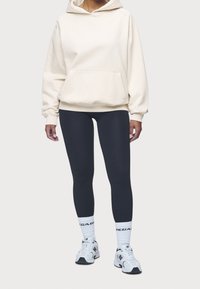 Sudadera con capucha color crema con bolsillo frontal, combinada con leggings negros, calcetines blancos con letras y zapatillas grises con detalles en negro.