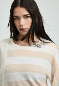 Junge Frau mit langen dunklen Haaren, die einen beige-weißen gestreiften Pullover trägt, schaut direkt mit einem neutralen Ausdruck, hellgrau im Hintergrund.