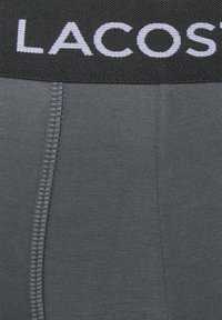 Grijze boxershorts hebben een zwarte elastische tailleband met "LACOSTE" in witte letters. De stof heeft een gladde, elastische textuur.