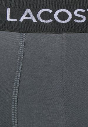 Des boxer-briefs gris avec une ceinture élastique noire sur laquelle est inscrit "LACOSTE" en lettres blanches. Le tissu présente une texture lisse et extensible.