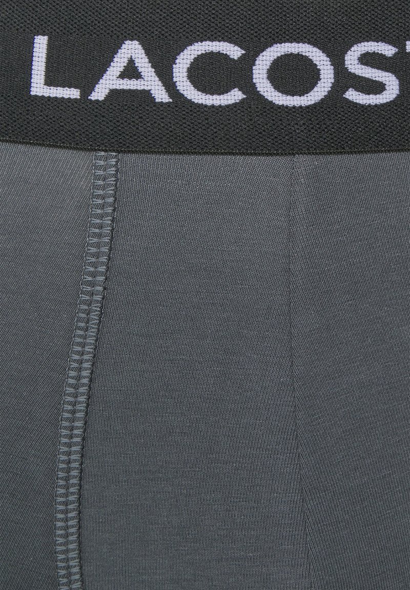 Grijze boxershorts hebben een zwarte elastische tailleband met "LACOSTE" in witte letters. De stof heeft een gladde, elastische textuur.