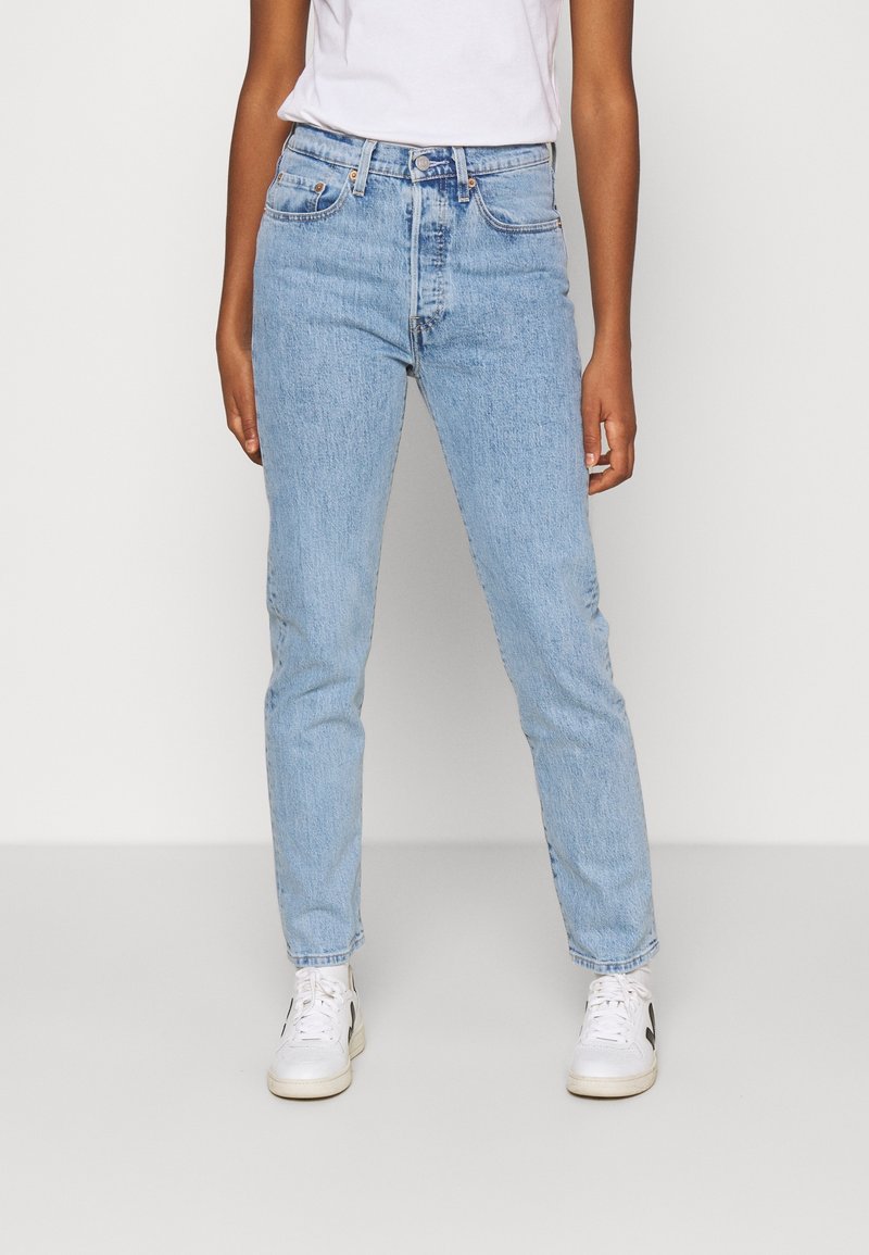 Personne portant un jean bleu clair à jambes droites, des baskets blanches et un t-shirt blanc, debout devant un fond uni.