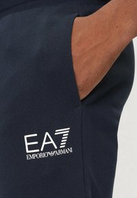 Marina blå träningsbyxor i mjukt tyg, med en sidficka och vitt EA7 Emporio Armani-logotyp tryckt på låret.