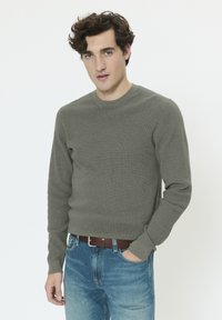 Pull vert en maille avec un col rond, manches longues et ourlet côtelé, assorti à un jean en denim bleu clair et une ceinture en cuir marron.