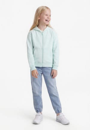 Lachend meisje met lang blond haar draagt een lichtblauwe hoodie met ritssluiting, lichtblauwe jogger jeans en witte sneakers, staand op een witte achtergrond.