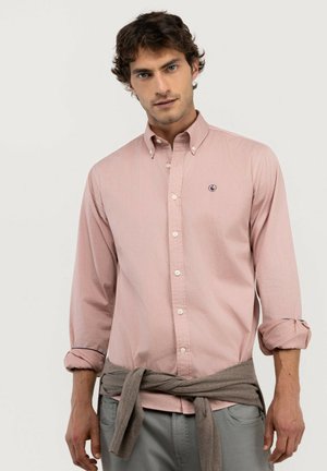 Hombre con cabello rizado y oscuro que lleva una camisa rosa claro abotonada con las mangas remangadas y un suéter gris atado alrededor de su cintura.