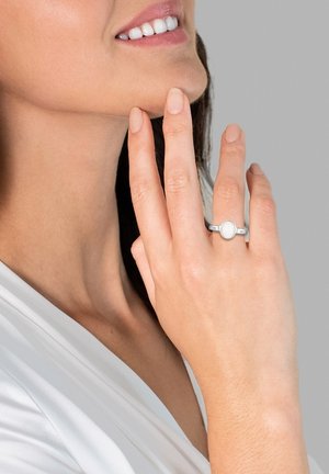 Silberner Ring mit einem ovalen weißen Stein, am Handgelenk getragen. Glatte Metallband mit polierter Oberfläche. Neutrale Hautfarbe, helle Nagelfarbe.