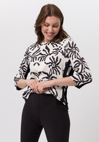 Witte blouse met gedurfde zwarte bloemenpatronen, driekwart mouwen en contrasterende zwarte zijpanelen. Zachte textuur, relaxte pasvorm.