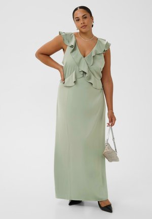 Femme portant une robe maxi vert clair sans manches avec des volants, tenant un petit sac beige et portant des chaussures noires pointues.