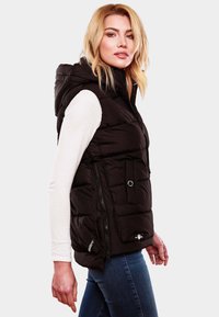 Gilet matelassé noir avec capuche, fermeture éclair à l'avant, poches latérales, texture matelassée et logo au niveau de l'ourlet, porté sur une chemise blanche à manches longues.