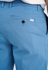 Pantaloni azzurri chiari realizzati in tessuto morbido, dotati di una tasca posteriore con un bottone nero e un piccolo etichetta con il nome del marchio.