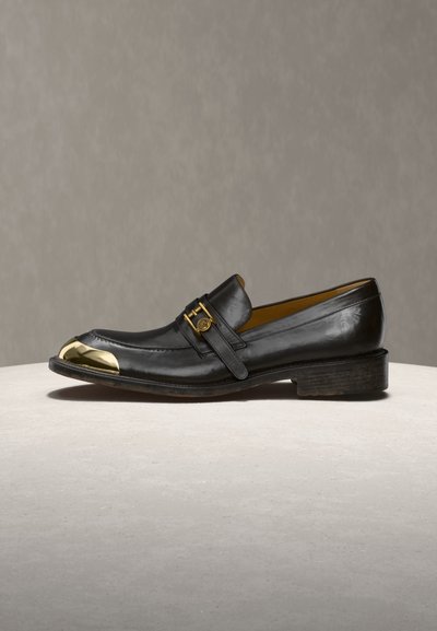Μαύρο δερμάτινο μοκασίνι με χρυσή μεταλλική μύτη και αντίστοιχο χρυσό πόμολο στο πλάι, εκτίθεται σε ανοιχτή επιφάνεια με ουδέτερο φόντο.