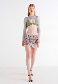 Groene bikini-top met een uitsnijdontwerp, gecombineerd met een korte rok met zebra-strepen en bloemmotieven. Lange, doorzichtige grijze mouwen. Zilveren sandalen.