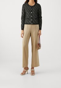 Cardigan noir à motif avec col rond et boutons, associé à un pantalon ample beige et des talons ouverts crème. Petit sac à main dans la main.