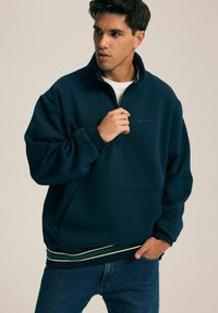 Granatowy polarowy sweatshirt z półzamkiem, z prążkowanym dołem w beżowe i zielone paski oraz z kieszenią na przodzie. Posiada subtelną grafikę na klatce piersiowej.