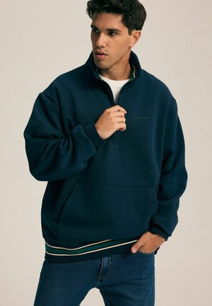 Granatowy polarowy sweatshirt z półzamkiem, z prążkowanym dołem w beżowe i zielone paski oraz z kieszenią na przodzie. Posiada subtelną grafikę na klatce piersiowej.