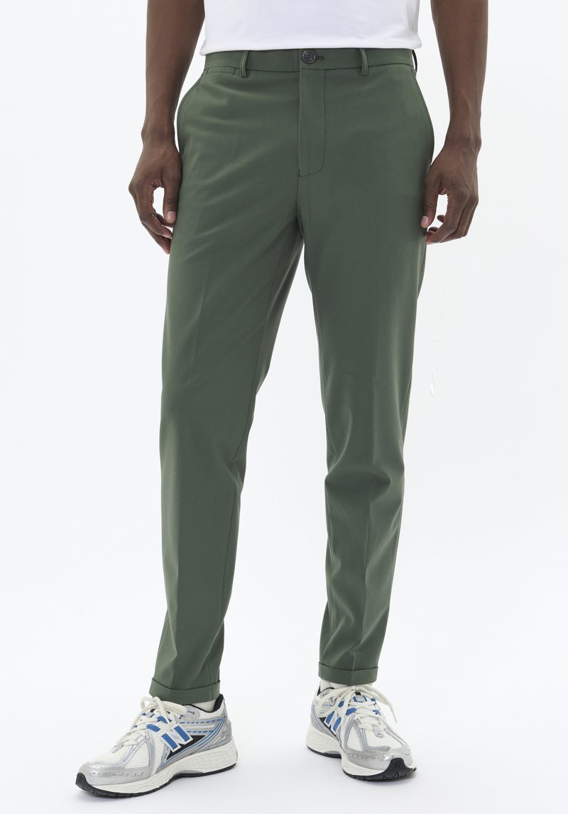 Pantalones verdes ajustados de tela suave, con delante plano, presillas para el cinturón y dobladillos rematados, combinados con zapatillas de running gris claro.