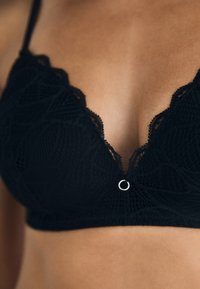 Svart blonde bralette med dyp utringning, med kompliserte blomstermønstre og en sølvringdetalj i midten.