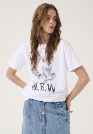 T-shirt blanc en coton avec un motif d'aigle imprimé noir et le texte "M.E.W.". Porté avec une jupe en jean bleu, les mains placées sur les hanches.