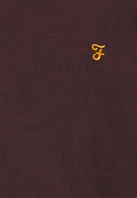 Bordowa koszulka z bawełny, z małym, żółtym, haftowanym logo „F” z przodu. Miękka tekstura, casualowy design.
