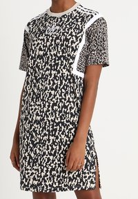 Šaty s krátkým rukávem s černobéžovým abstraktním potiskem, bílé akcenty na ramenech, oblečené osobou stojící s uvolněnou pravou rukou.