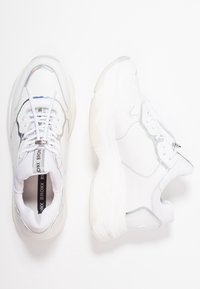Un par de zapatillas deportivas blancas y gruesas con detalles iridiscentes, cordones blancos, plantilla de marca y suelas de goma gruesas sobre un fondo blanco.