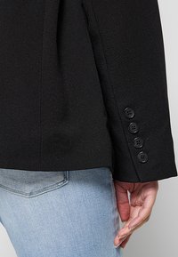Blazer negro a medida con diseño estructurado, que presenta una textura suave y cuatro botones negros en la manga, combinado con jeans azul claro.