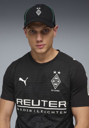 Młody mężczyzna w czarnej czapce Borussia Mönchengladbach i czarnej koszulce Puma z logo klubu i napisem sponsora Reuter.
